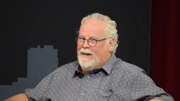 Author Michael Connelly, with white hair and a white beard, wearing glasses and a gray patterned shirt, smiling on a stage
