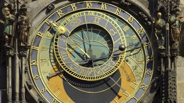 An ornate gold and blue astronomical clock face with Roman numerals, an inner ring of zodiac symbols and carved figures on either side