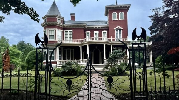 A red Victorian house with a tower and a white porch, seen through a black wrought-iron gate topped with bats and patterned with spider webs